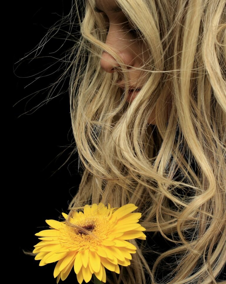 Blonde woman with flowing hair holding a vibrant yellow flower against a dark background.