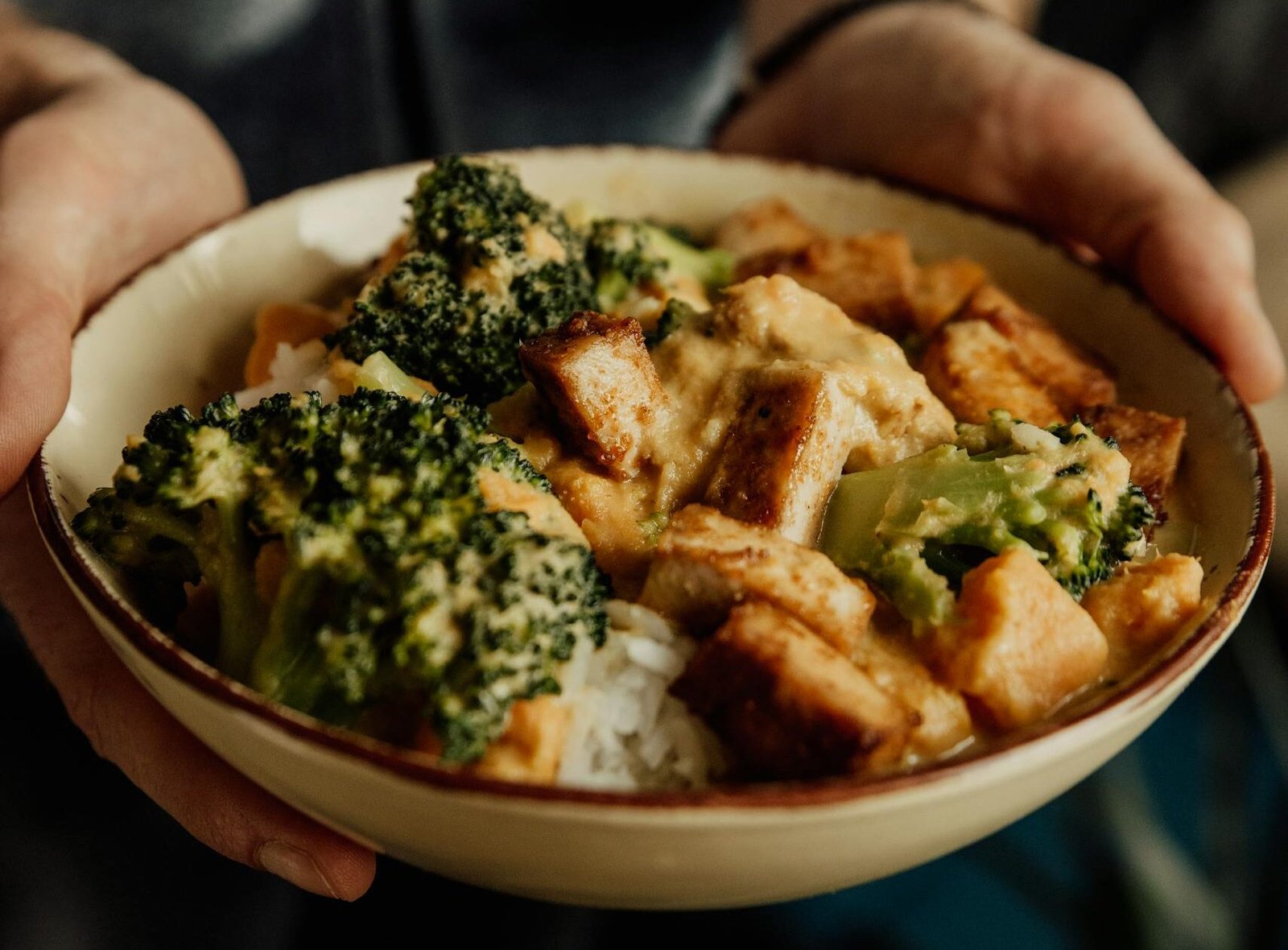 Close-up of a person holding a bowl of broccoli, tofu, and rice, offering a nutritious meal option.
