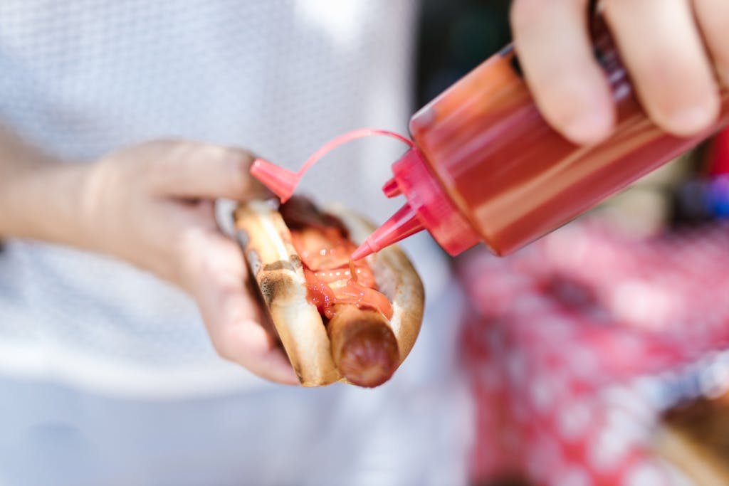 Person adding ketchup to a grilled hot dog held in hands outdoors.