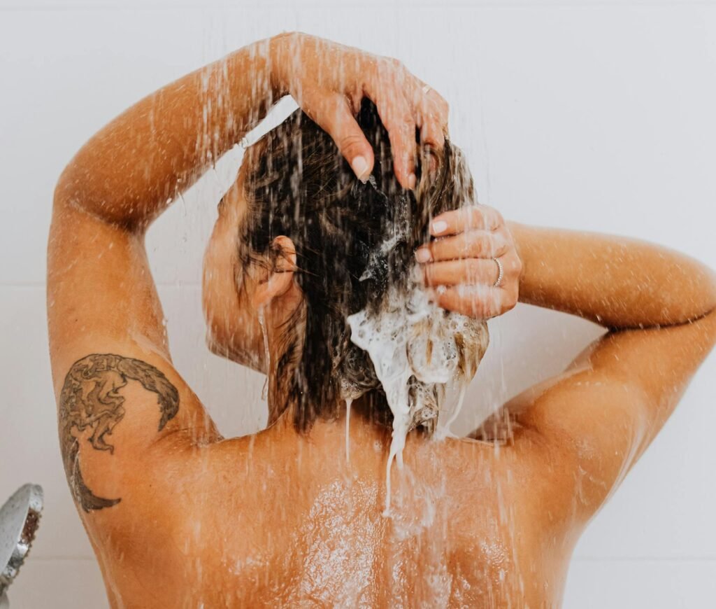 Rear view of a woman washing her hair in the shower, showcasing a tattoo and emphasizing hygiene.