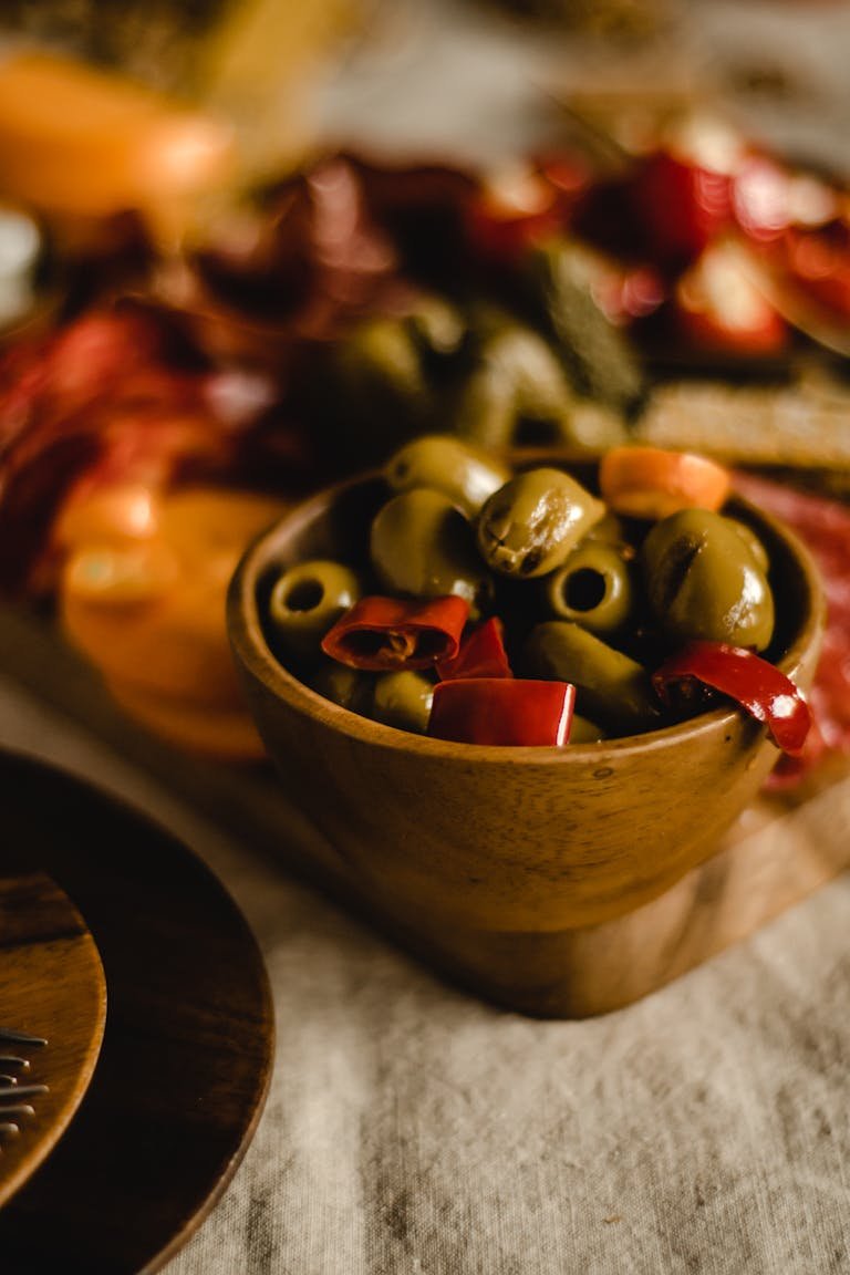 Close-up of a rustic wooden bowl filled with fresh olives and peppers in a cozy setting.