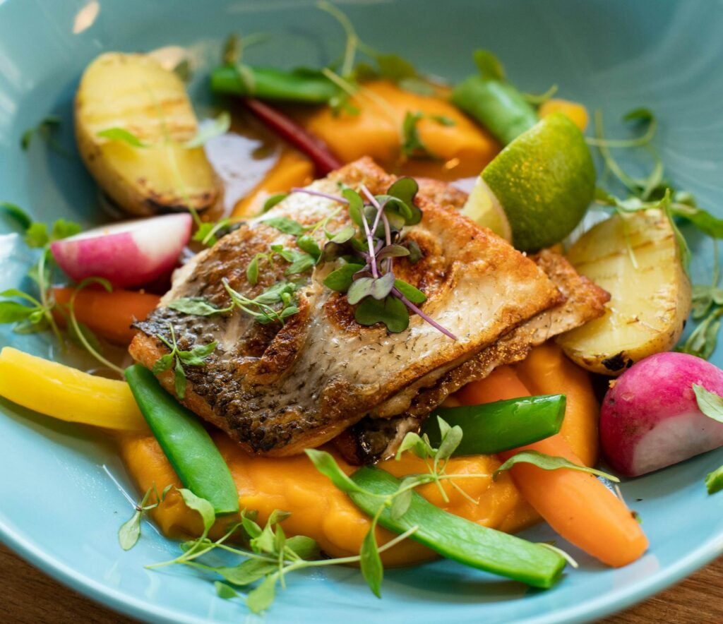 Colorful dish featuring grilled fish, vegetables, and microgreens in a blue bowl.